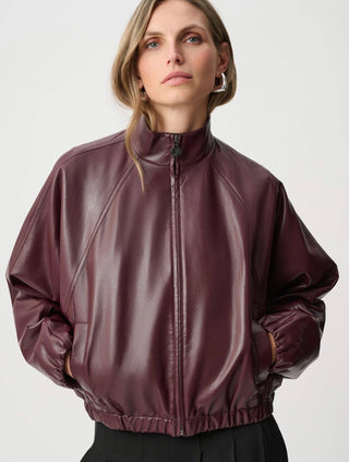 A woman wearing a glossy, cropped bomber jacket in a deep burgundy or plum color, paired with black trousers, against a plain light gray studio background.